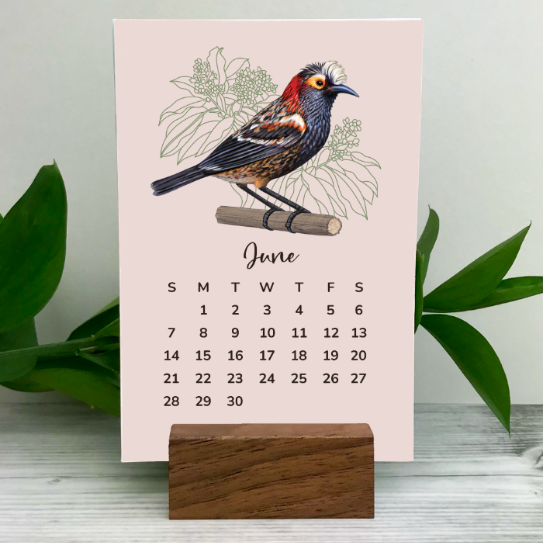 Desk calendar with a Hawaiian bird illustration on a pink background, standing on a wooden block with green leaves in the background.