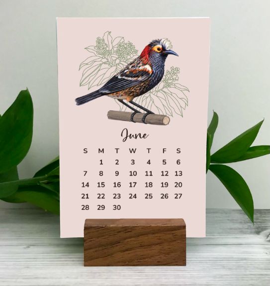 Desk calendar with a Hawaiian bird illustration on a pink background, standing on a wooden block with green leaves in the background.